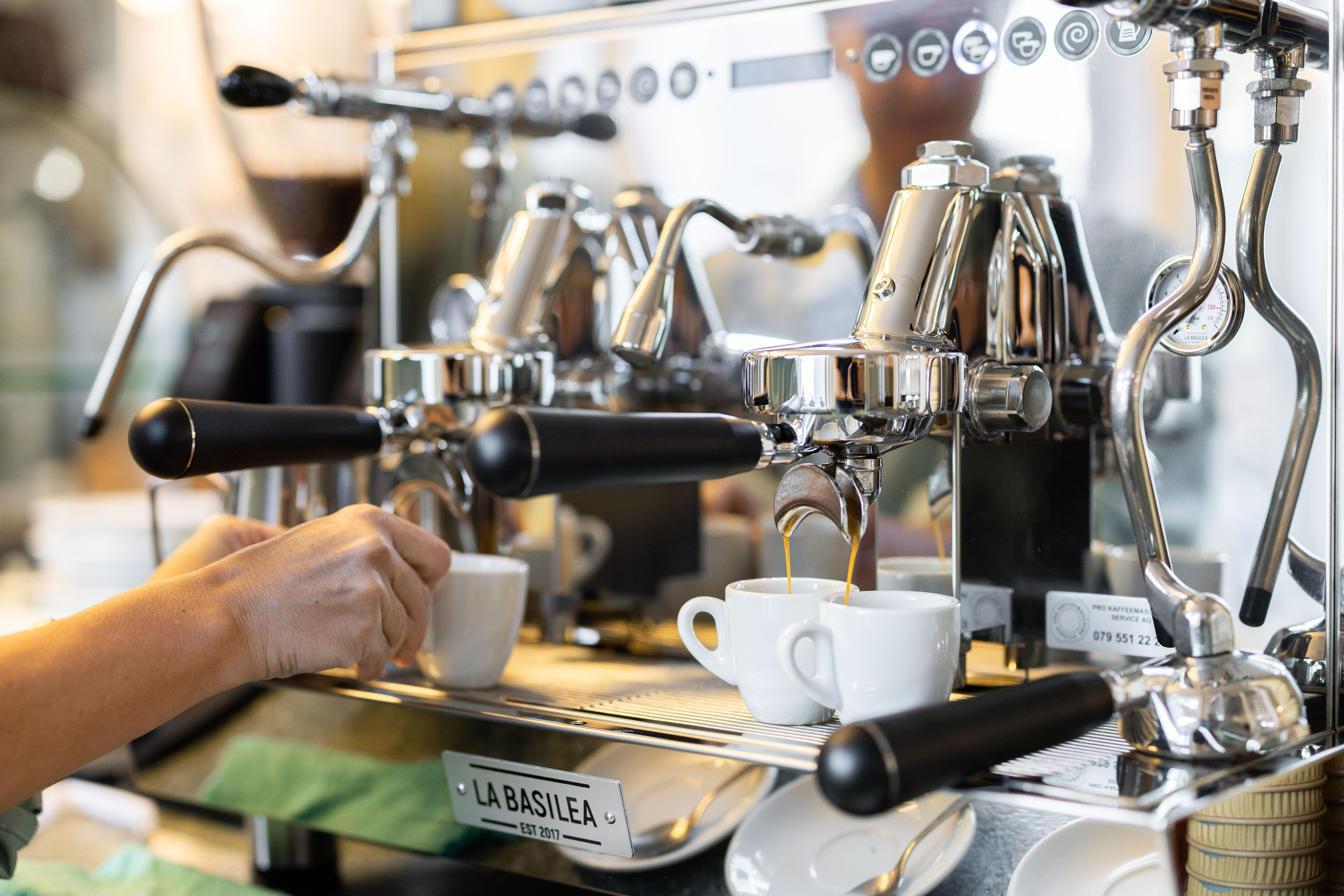 Eine Person stellt eine Tasse unter den Kolben einer Industriekaffeemaschine, während daneben zwei Kaffees gebrüht werden.