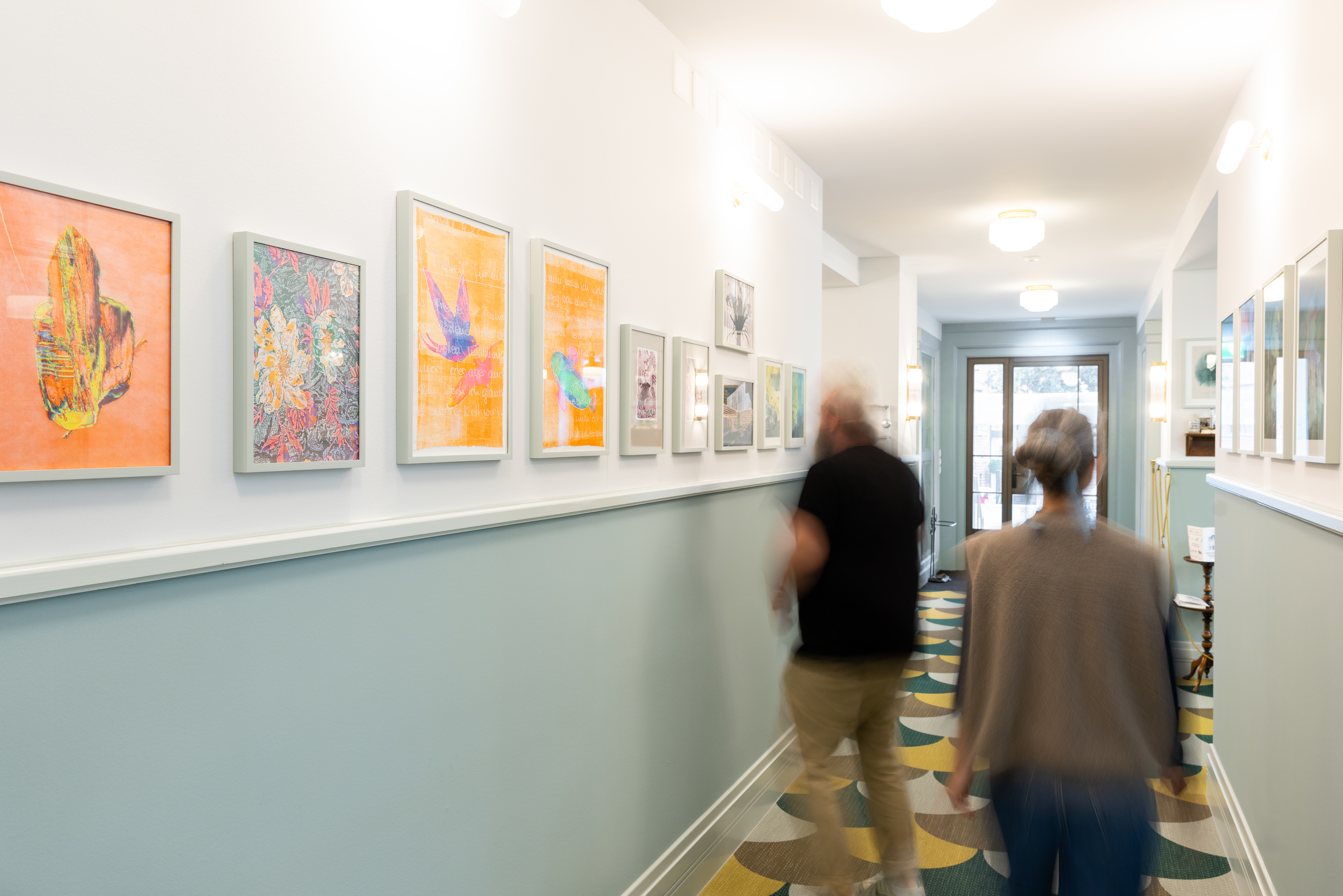 Eine Wechselausstellung von Bildern im Socin Haus. Künstlerin: Linda Heydegger. Schräge Sicht auf zwei laufende Menschen vor den Bildern der Ausstellung.